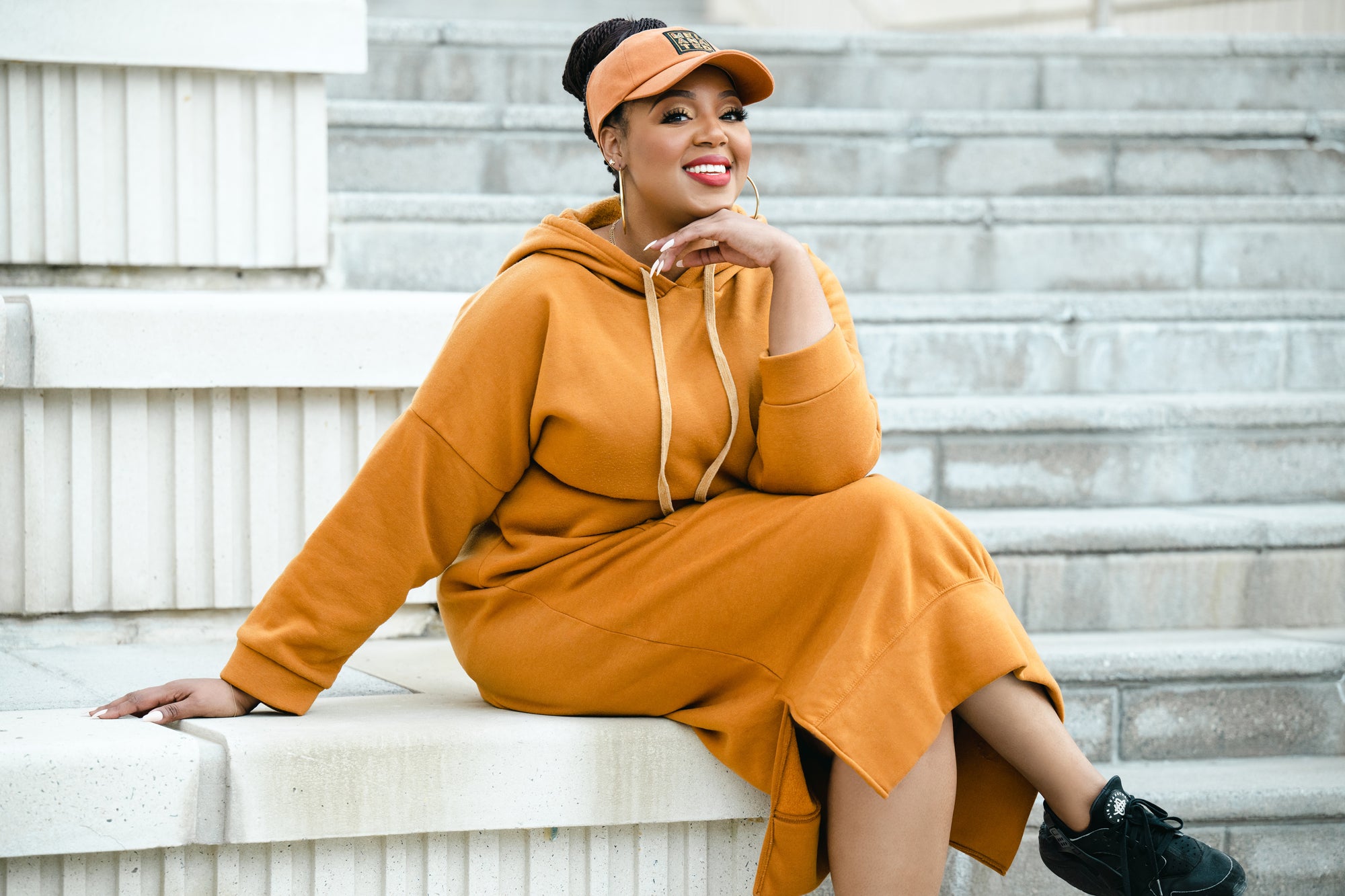 Jasmine Hudson Co-Founder of Black Paper Party sitting on stairs wearing a tan hoodie dress and tan visor