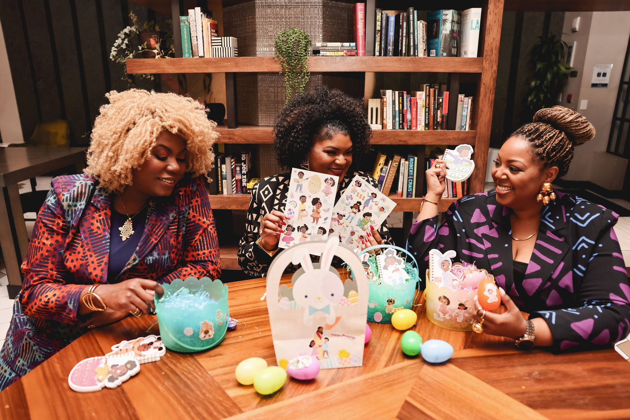 "Three women sitting around a table, smiling and engaging with each other, surrounded by colorful party supplies and decorations from Black Paper Party. They are examining products and appear to be having a joyful time. The atmosphere is lively and celebrates cultural diversity.