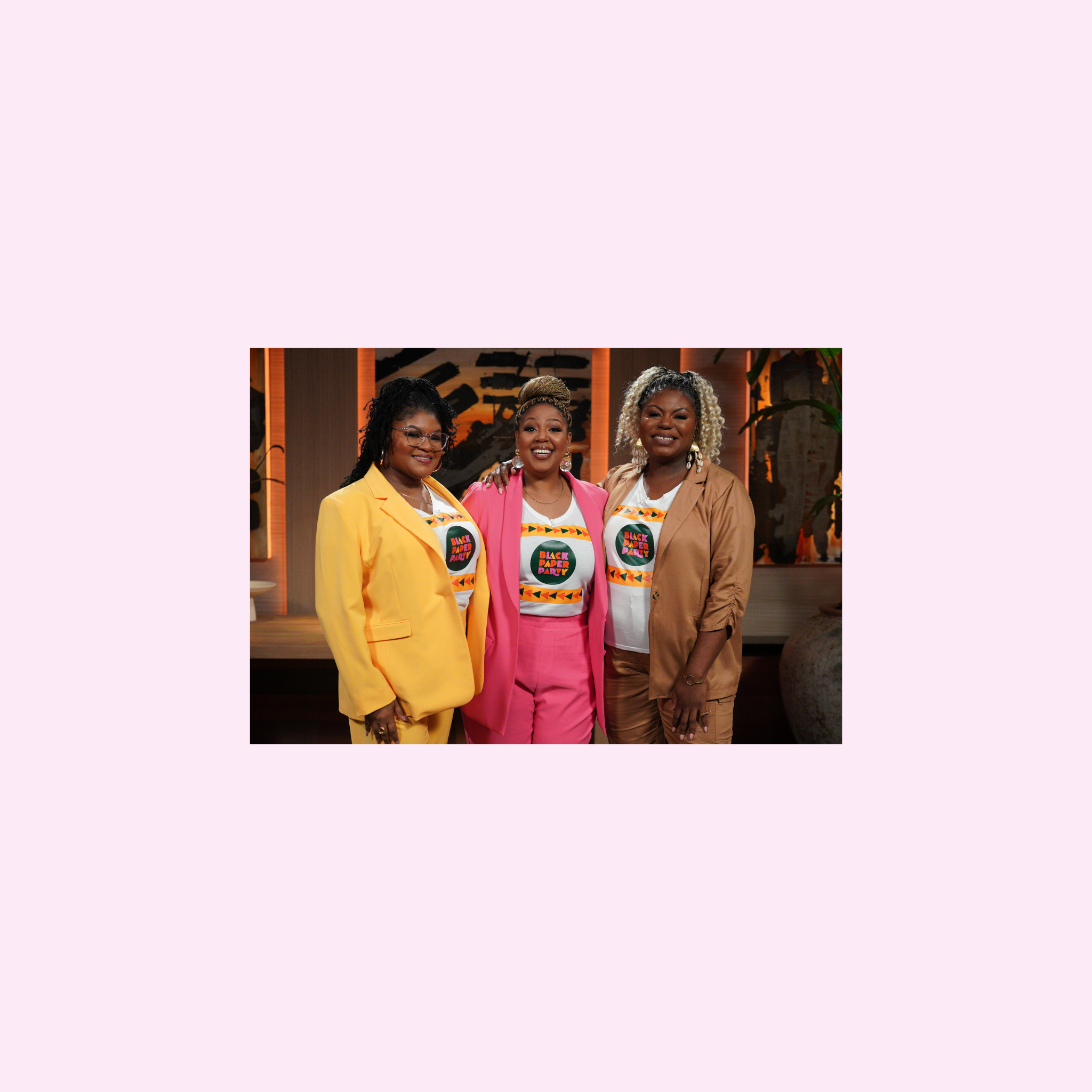 Three women are standing side-by-side, smiling, in a warmly lit room. The woman on the left wears a yellow blazer, the middle woman is in a pink jumpsuit, and the one on the right dons a beige suit. All three are sporting colorful T-shirts with a vibrant graphic design.