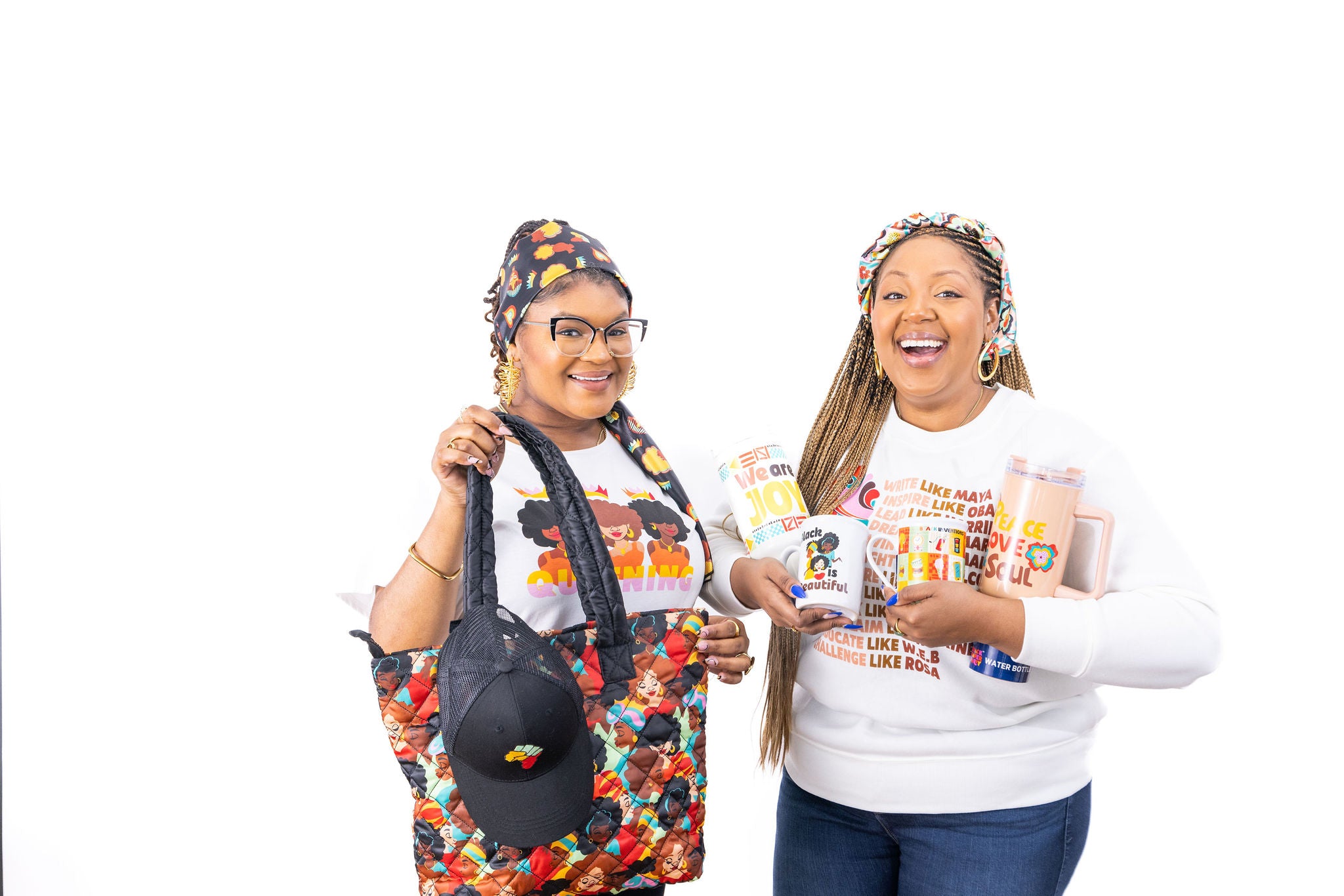 The image shows two women smiling brightly against a white background. The woman on the left is wearing glasses and a colorful head wrap, holding a patterned bag and a black cap. The woman on the right, with braided hair and a headband, is holding a clear tumbler and a mug with inspirational quotes. Both wear white shirts with a joyful print, exuding a positive and spirited vibe.