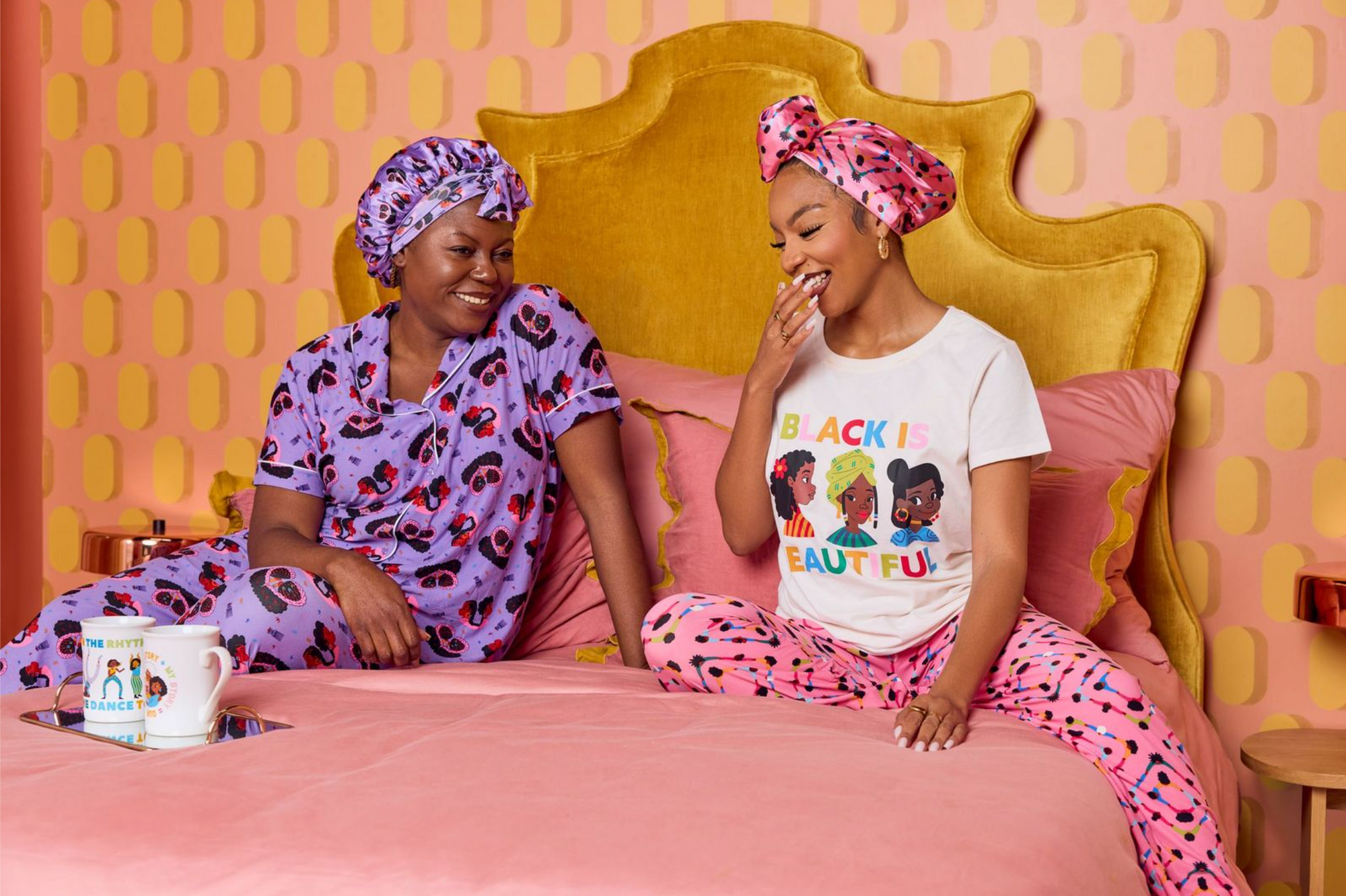 Two Black women in colorful pajamas laugh together on a pink and gold bed, surrounded by cozy textures, bold patterns, and playful mugs — capturing the everyday joy and warmth of Black Paper Party.