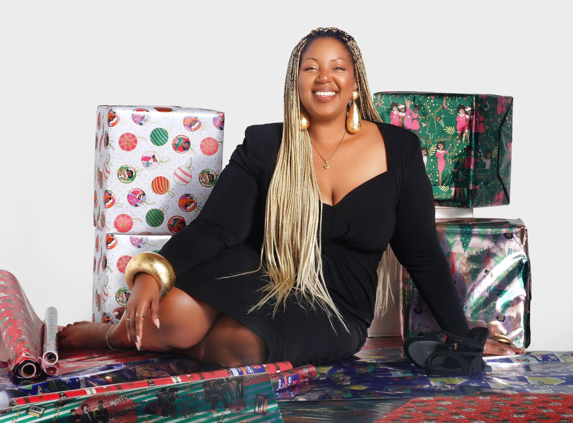 A woman wearing a black dress sits confidently on a white cube, surrounded by festive gift-wrapped boxes and vibrant rolls of Black Paper Party wrapping paper. She smiles at the camera. The scene teases the upcoming holiday season with bold colors, Black representation, and stylish celebration.