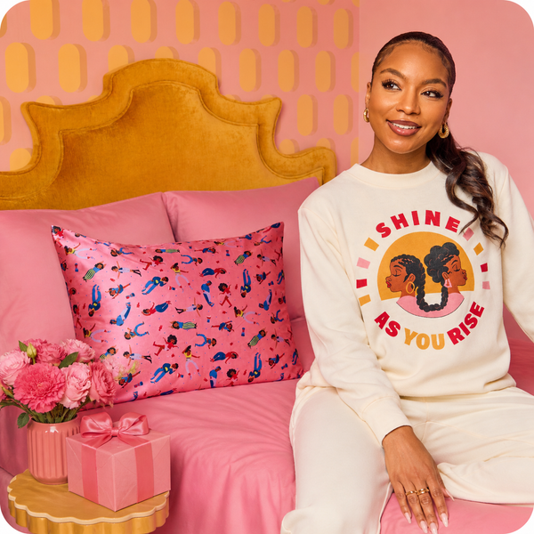 Woman sitting on a pink bed with a 'Shine As You Rise' sweatshirt, surrounded by decorative items.