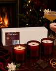 Three lit Noire Noel candles styled beside a fireplace with cocktails.